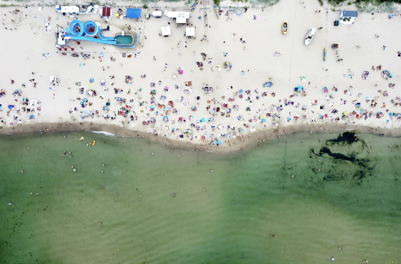 Plaża w Stegnie Plaża pełna ludzi wypoczywających na ręcznikach i pod parasolami, sztuczna zjeżdżalnia wodna w lewym górnym rogu, łódki oraz jasny piasek przechodzący w zielonkawą wodę, zauważalna ciemna plama w wodzie.