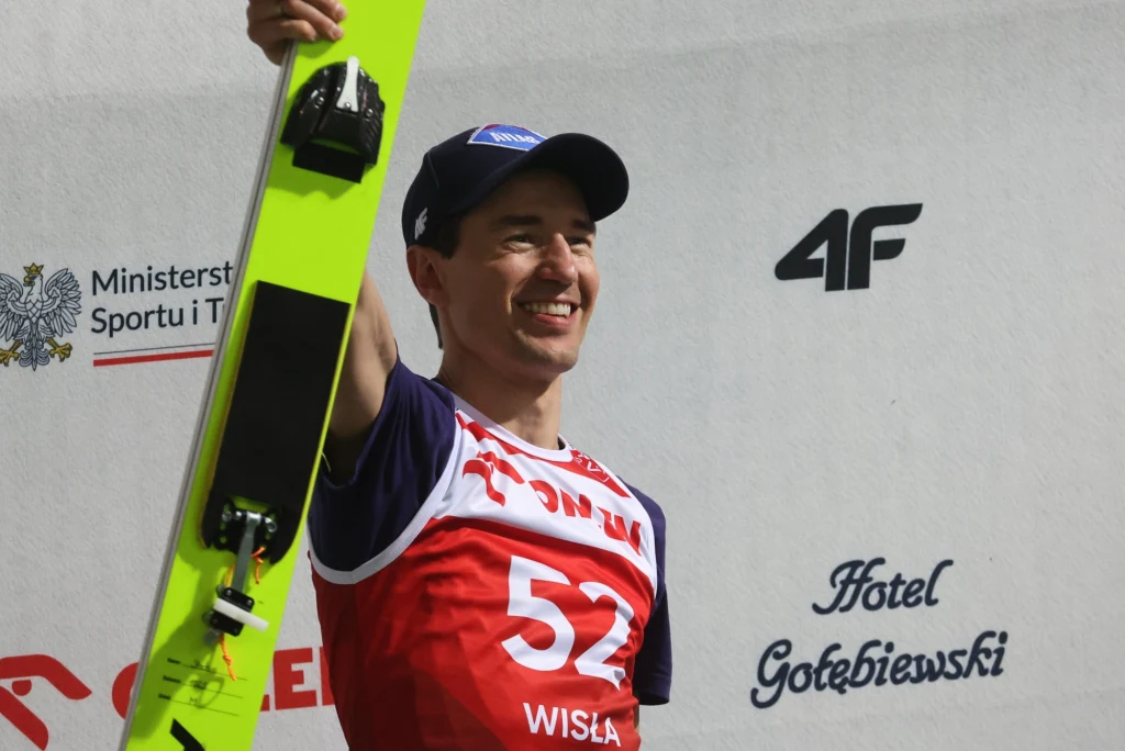 Kamil Stoch Skoczek narciarski w stroju sportowym, trzymający narty w uniesionej ręce, uśmiechnięty, w tle widoczne logotypy sponsorów i napisy związane z miejscem wydarzenia.