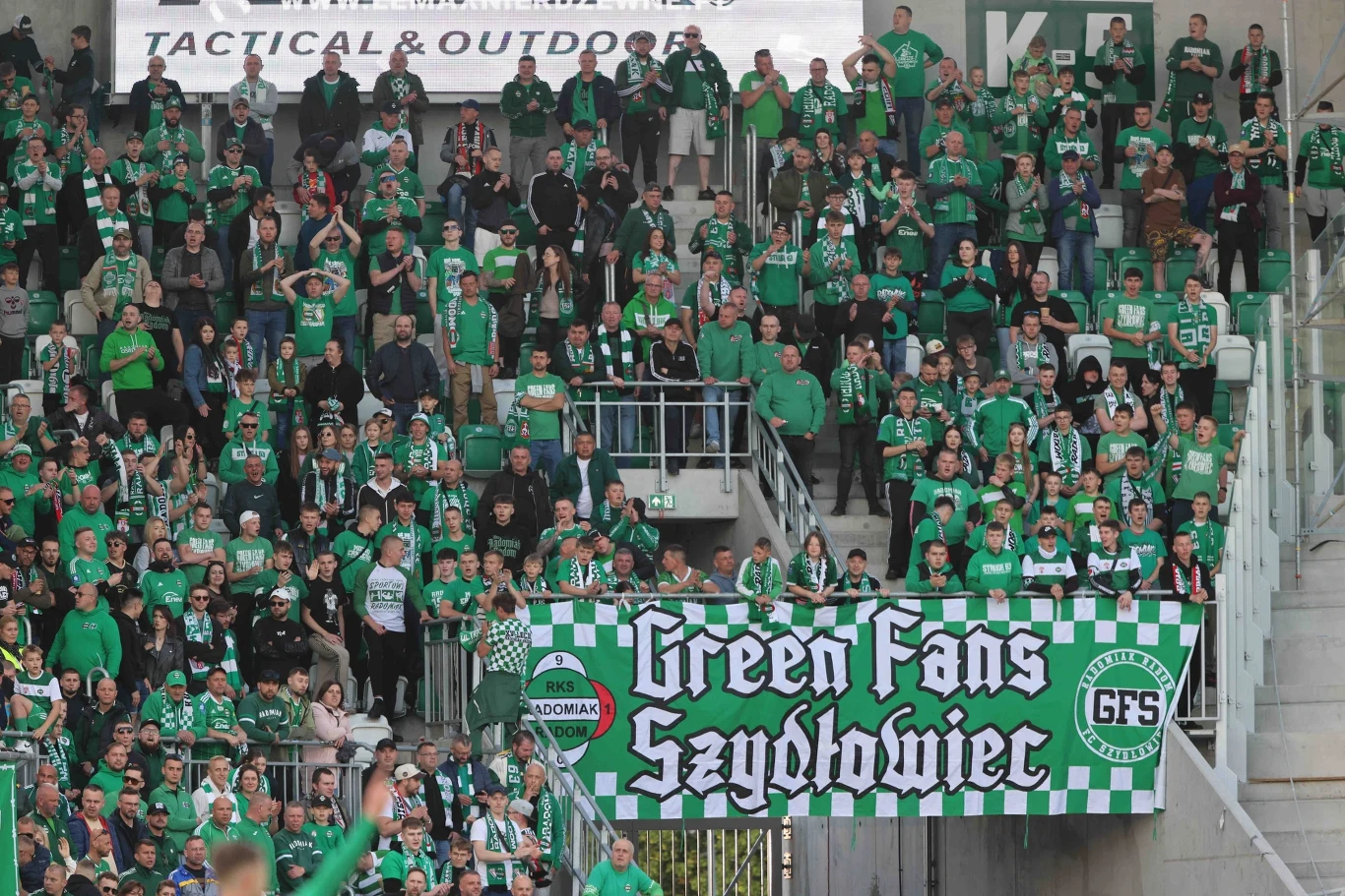 Tłum kibiców ubranych w zielone barwy klubowe zgromadzony na stadionie, wiele osób trzyma szaliki oraz transparent z napisem Green Fans Szydłowiec, atmosfera pełna entuzjazmu i wsparcia dla drużyny.