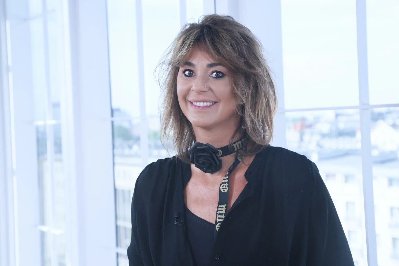 Kobieta o brązowych włosach ubrana w ciemną koszulę z uśmiechem na twarzy, na szyi zawieszony aparat fotograficzny, w tle jasne okna i rozmyty widok miasta.