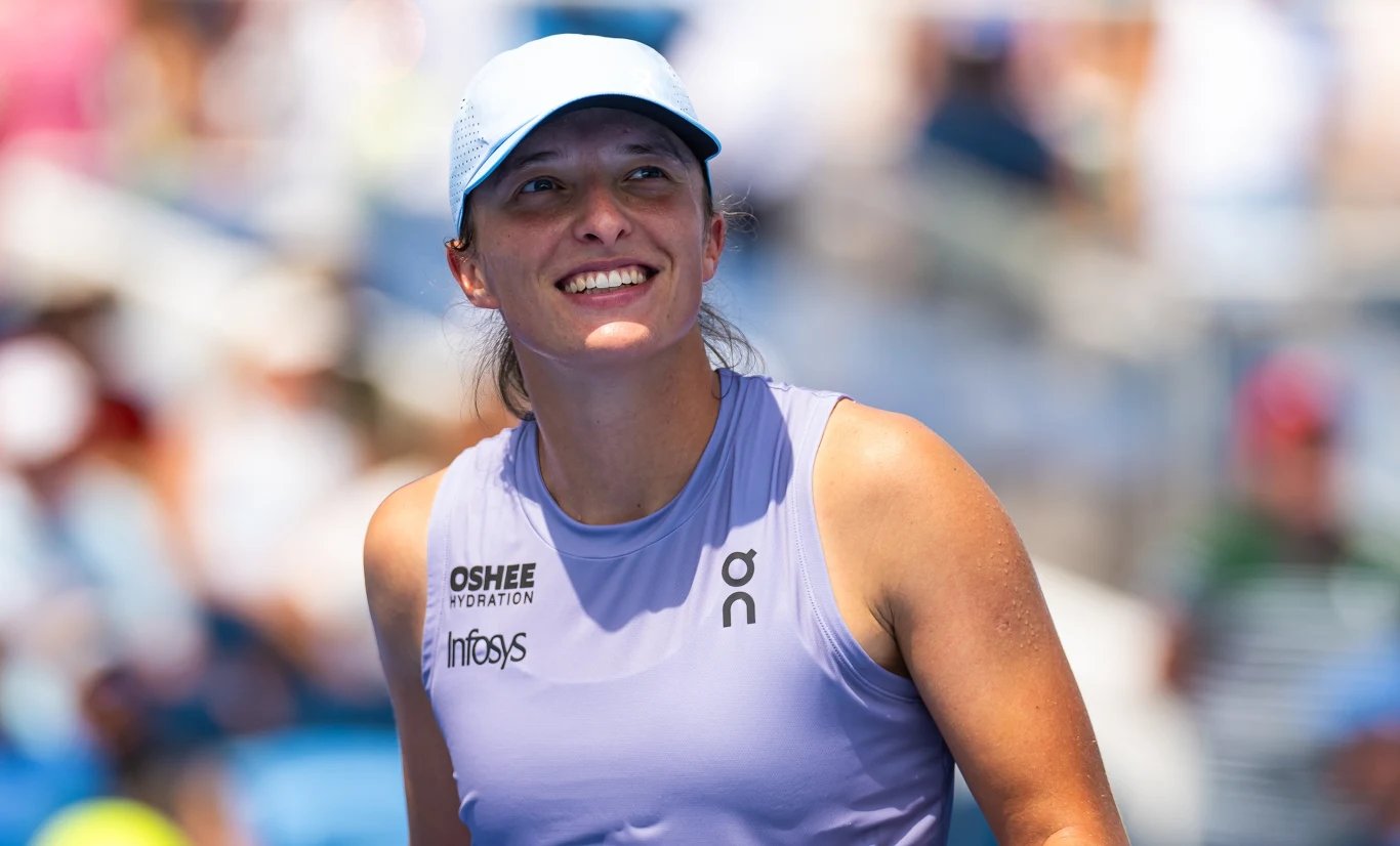 Iga Świątek rywalizowała z Jeleną Rybakiną o awans do finału WTA 1000 w Cincinnati Uśmiechnięta kobieta w sportowej koszulce i czapce z daszkiem na tle rozmytego stadionu tenisowego, promienie słońca oświetlają jej twarz.