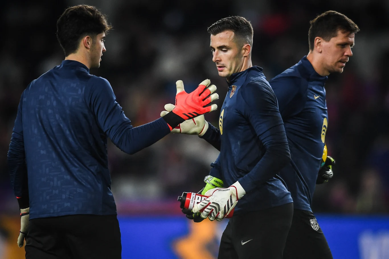 Trzech piłkarzy ubranych w granatowe stroje sportowe, dwóch z nich uderza się rękawicami w geście powitania lub wsparcia na boisku, w tle niewyraźna publiczność i trybuny stadionu.