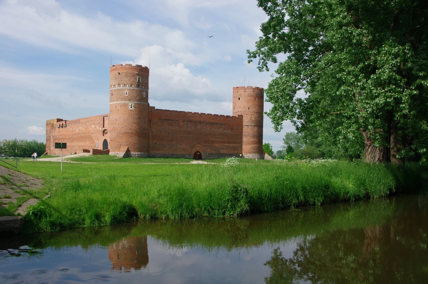 Ceglany zamek otoczony zielonym trawnikiem i dużymi drzewami, położony nad spokojnym zbiornikiem wodnym, odbijającym jego sylwetkę.