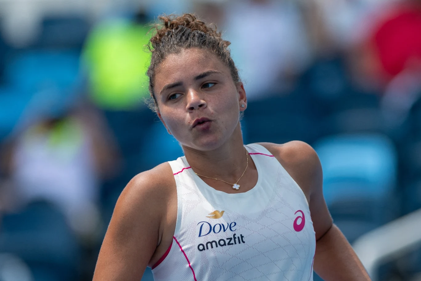 Młoda kobieta sportowiec w białym stroju tenisowym z widocznymi logotypami sponsorów, z intensywnym wyrazem twarzy, na tle rozmytych trybun stadionu sportowego.