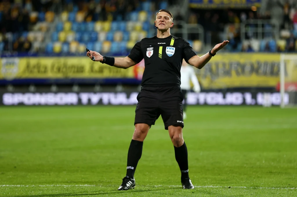 Sędzia piłkarski w czarnym stroju stoi na murawie stadionu z rozłożonymi rękami, w tle widoczne puste krzesełka na trybunach i nieczytelne transparenty.