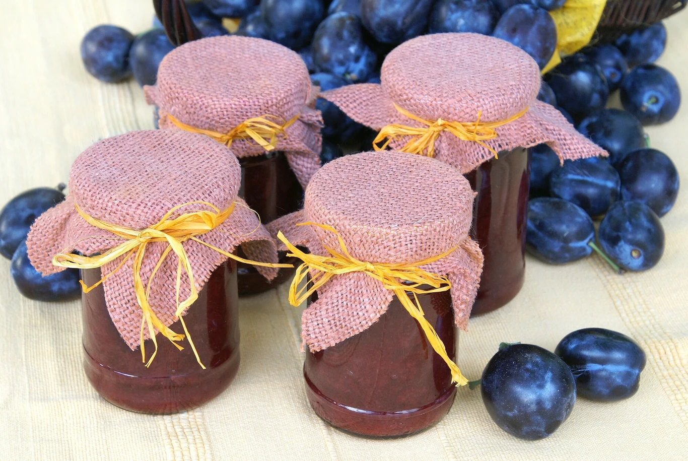 Cztery słoiki dżemu przykryte lnianą tkaniną i przewiązane żółtym sznurkiem, w tle świeże śliwki rozsypane na jasnym obrusie.