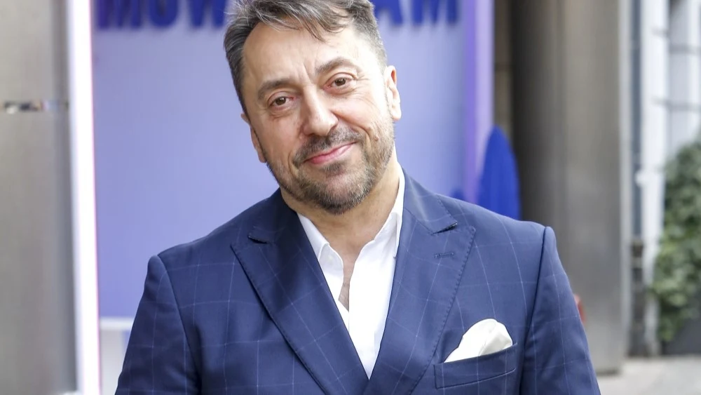 Marcin Miller Mężczyzna w średnim wieku w eleganckim, granatowym garniturze z białą poszetką, stojący na tle nowoczesnej zabudowy miejskiej, z delikatnym uśmiechem na twarzy.