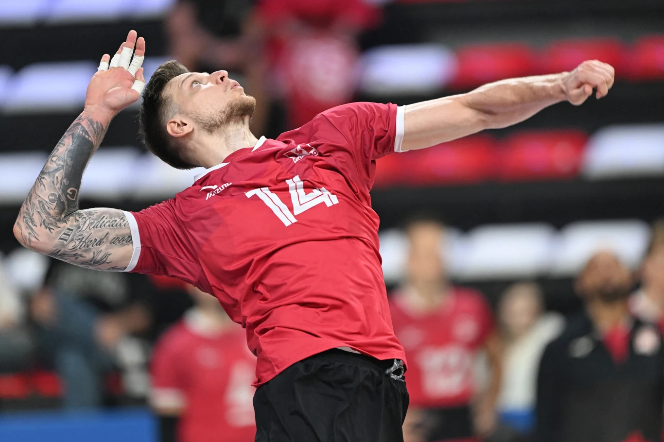 Sportowiec w czerwonym stroju z numerem 14 wykonuje dynamiczny ruch, najprawdopodobniej serwuje piłkę podczas meczu siatkówki, w tle widoczne rozmazane sylwetki publiczności oraz ławki w barwach czerwono-białych.