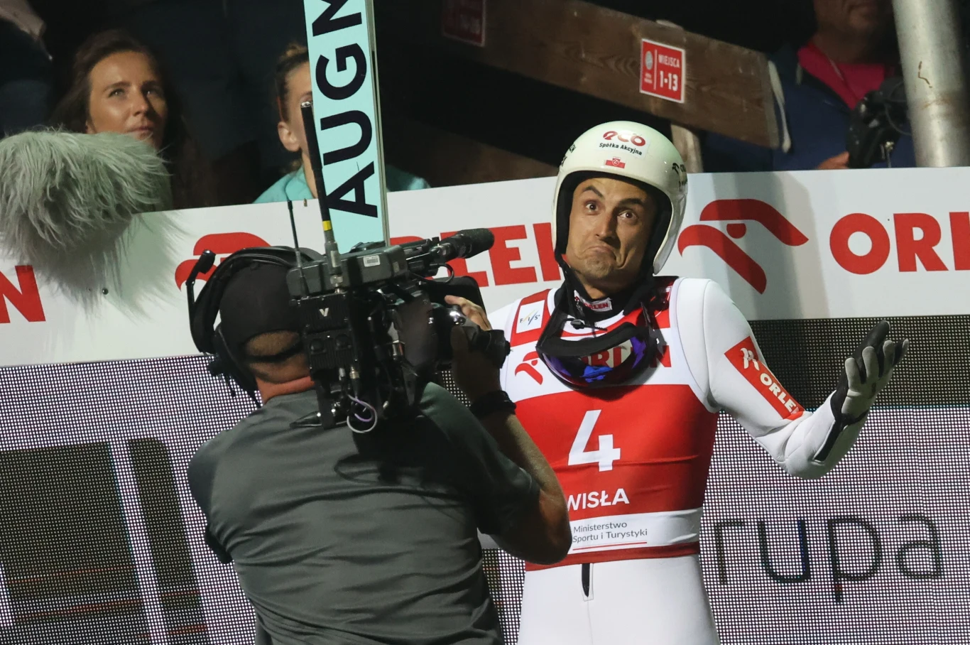 Maciej Kot Zawodnik skoków narciarskich w kombinezonie z numerem 4 gestykuluje przed kamerą telewizyjną podczas zawodów, z tyłu widoczne logo sponsora i publiczność.