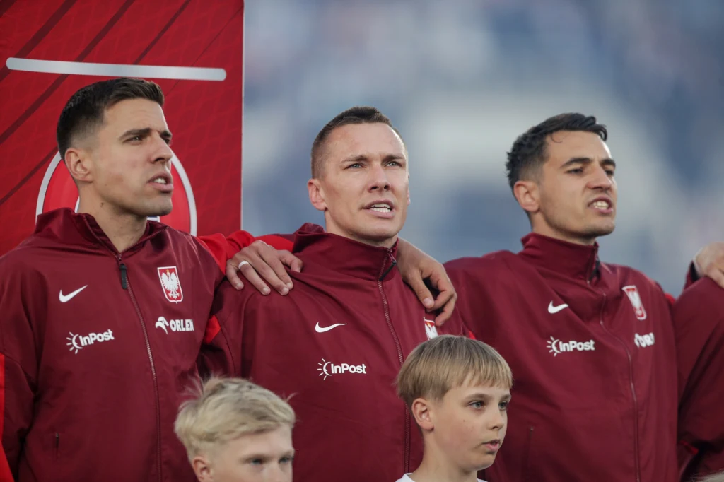 Jakub Kiwior (od prawej) Trzej piłkarze ubrani w bordowe bluzy reprezentacji Polski stoją razem z rękoma na ramionach, obok nich znajdują się dzieci ubrane na biało.