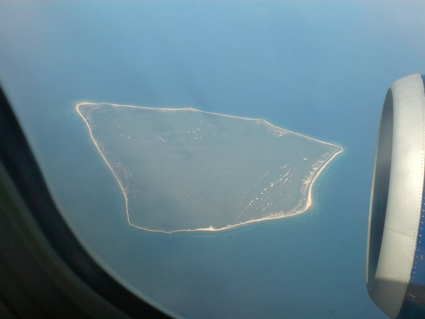 Widok wyspy otoczonej wodami morskimi, sfotografowanej z okna samolotu, z fragmentem silnika widocznym w prawym dolnym rogu kadru.