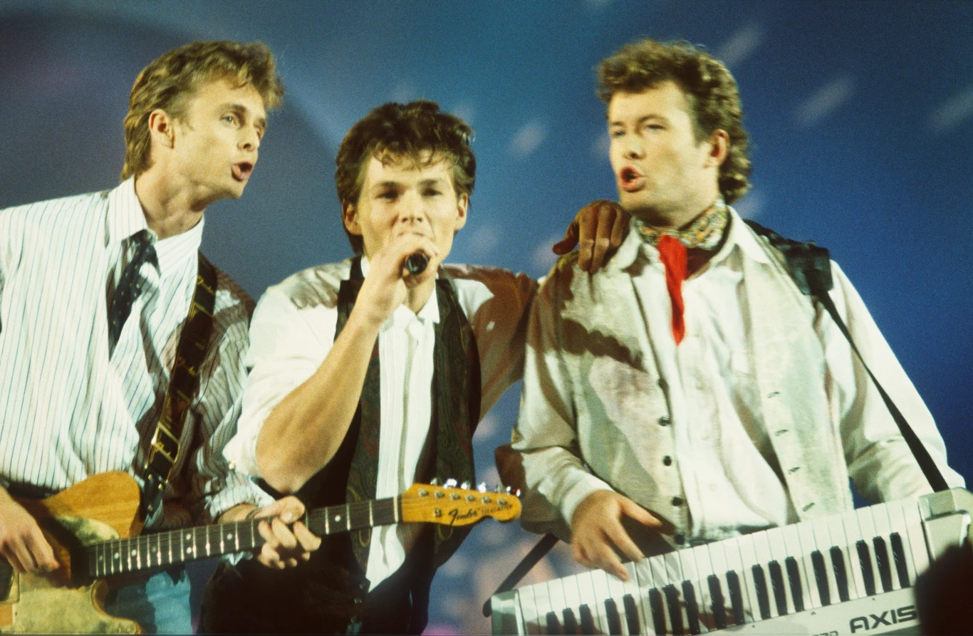 Zespół a-ha Trzech mężczyzn na scenie muzycznej, jeden śpiewa do mikrofonu, drugi gra na gitarze elektrycznej, a trzeci na klawiszach, wszyscy występują w jasnych koszulach, dynamiczne światła w tle tworzą koncertową atmosferę.