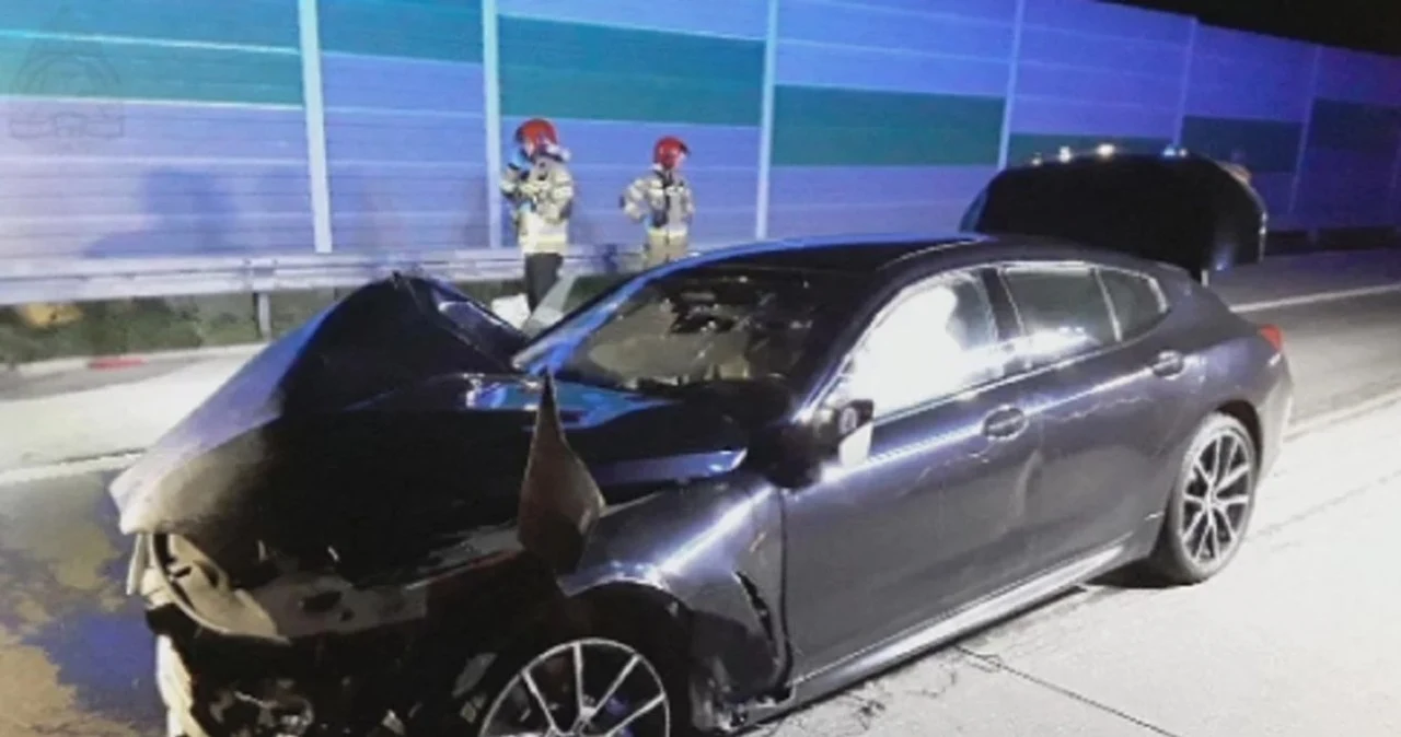 Tragiczny wypadek na autostradzie A1. BMW Sebastiana M. Rozbity samochód osobowy stoi na pasie ruchu, widoczne poważne uszkodzenia przodu pojazdu, w tle dwóch strażaków w umundurowaniu oraz osłony akustyczne przy drodze.