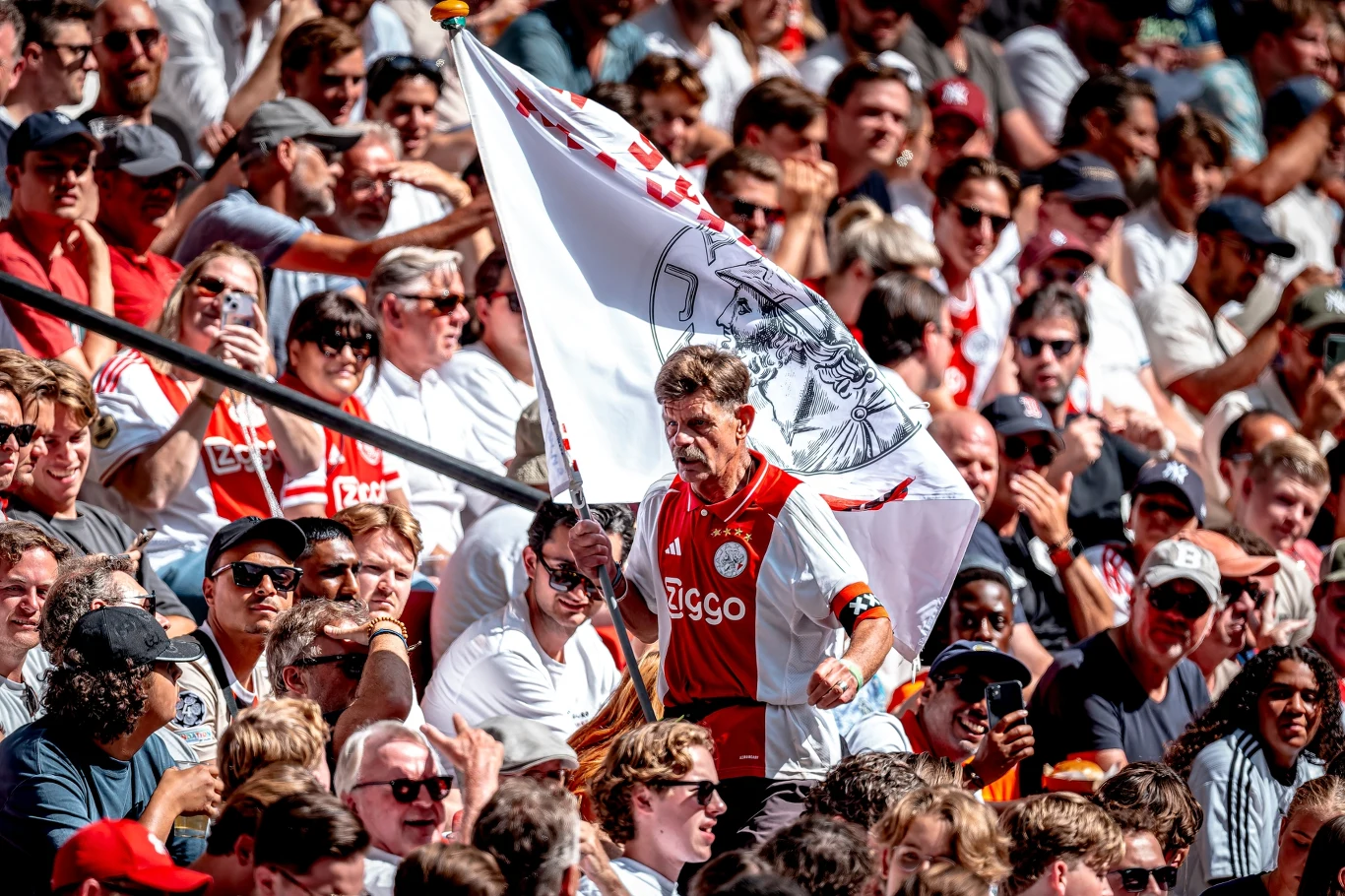 Kibice Ajaksu Amsterdam (zdj. ilustracyjne) Kibic w barwach klubu Ajax Amsterdam w tłumie fanów trzyma dużą flagę z klubowym motywem, wokół niego liczna grupa ludzi ubrana w stroje klubowe i codzienne, atmosfera trybun podczas meczu piłkarskiego.