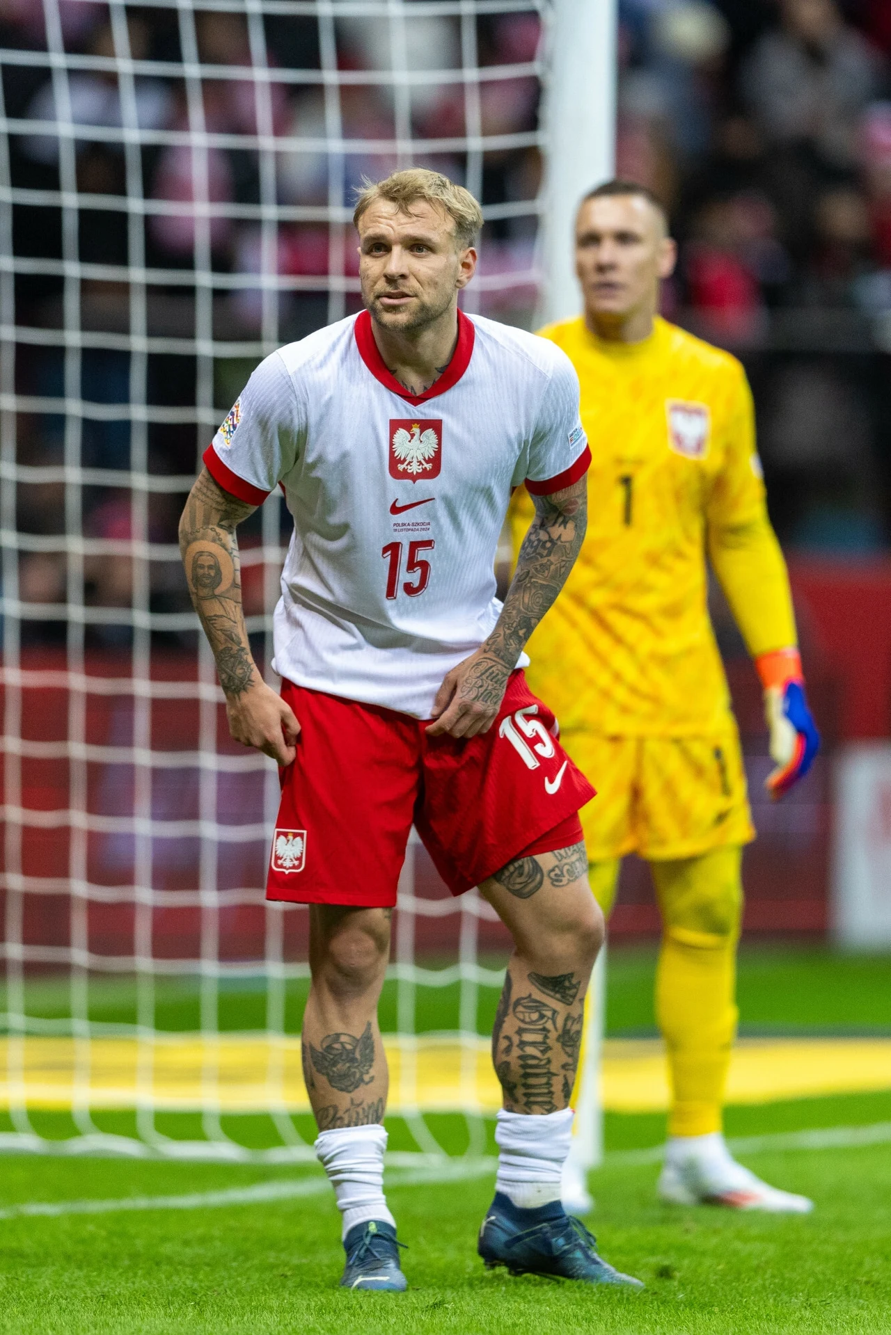 Tymoteusz Puchacz Piłkarz w białej koszulce z numerem 15 i czerwonych spodenkach z widocznymi tatuażami stoi na murawie, na drugim planie znajduje się bramkarz w żółtym stroju, w tle bramka i widownia stadionowa.