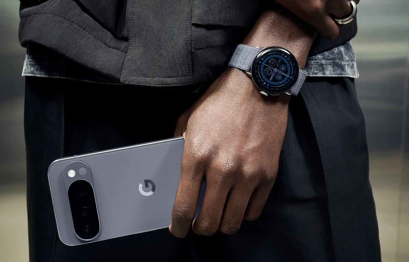 Google Pixel Watch 4 zaprezentowany. Znamy polskie ceny smartwatchy. Ciemnobrązowa dłoń trzyma szary smartfon Google Pixel, na nadgarstku noszony jest nowoczesny smartwatch z cyfrową tarczą i szarym paskiem, osoba ubrana jest w ciemną kurtkę.