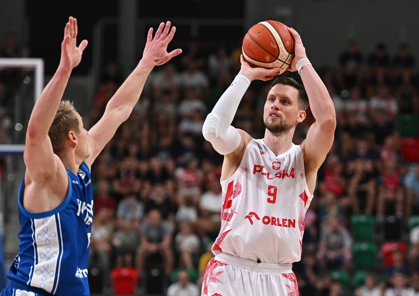 Mateusz Ponitka podczas meczu polskich koszykarzy z Finlandią Zawodnik reprezentacji Polski w białym stroju wykonuje rzut do kosza podczas meczu koszykówki, blokowany przez rywala z drużyny ubranej na niebiesko, w tle widownia na trybunach.