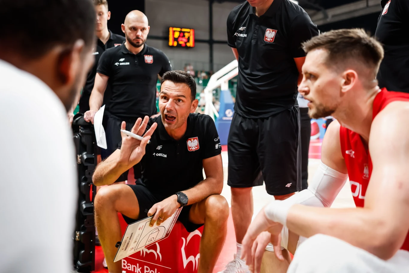 Trener Igor Milicic i koszykarze reprezentacji Polski w trakcie meczu Trener koszykówki w trakcie narady tłumaczy strategię drużynie zawodników ubranych w stroje reprezentacji Polski na hali sportowej, wokół zgromadzeni członkowie sztabu szkoleniowego.