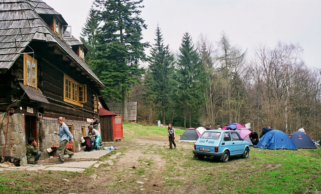 Maluchem pod namiot na trzy tygodnie? Czemu nie! Drewniany schronisko górskie otoczone lasem, przed budynkiem kilku ludzi rozmawia i odpoczywa, na trawiastym placu obok stoi niebieski fiat 126p, w tle rozstawione kolorowe namioty turystyczne, atmosfera biwaku i wypoczynku na łonie natury.