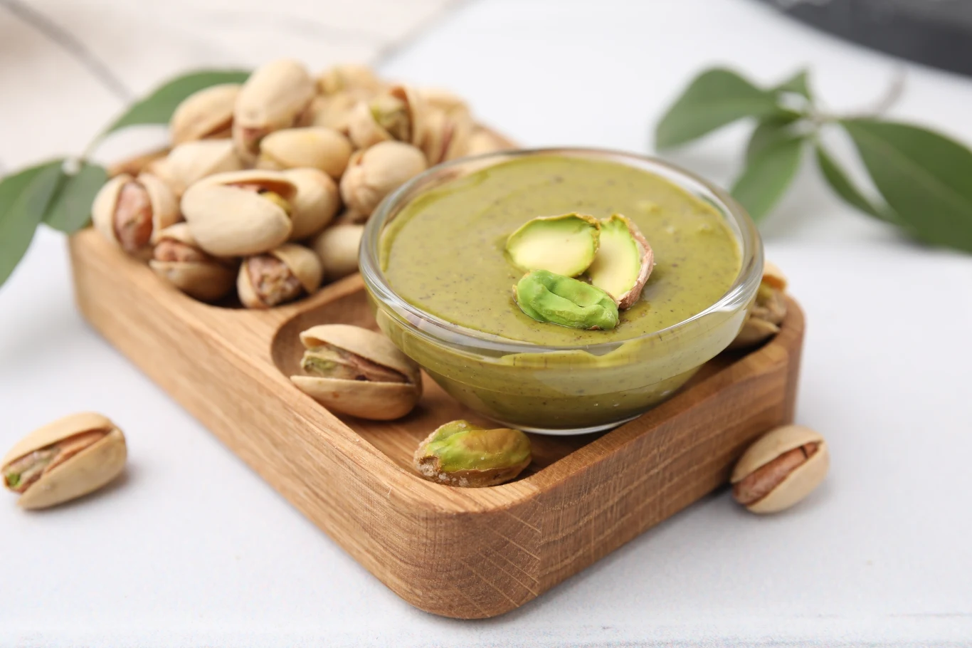 Szklana miseczka z kremem pistacjowym ozdobiona kawałkami pistacji, obok znajdują się całe, niełuskane pistacje na drewnianej podstawce, w tle zielony liść.