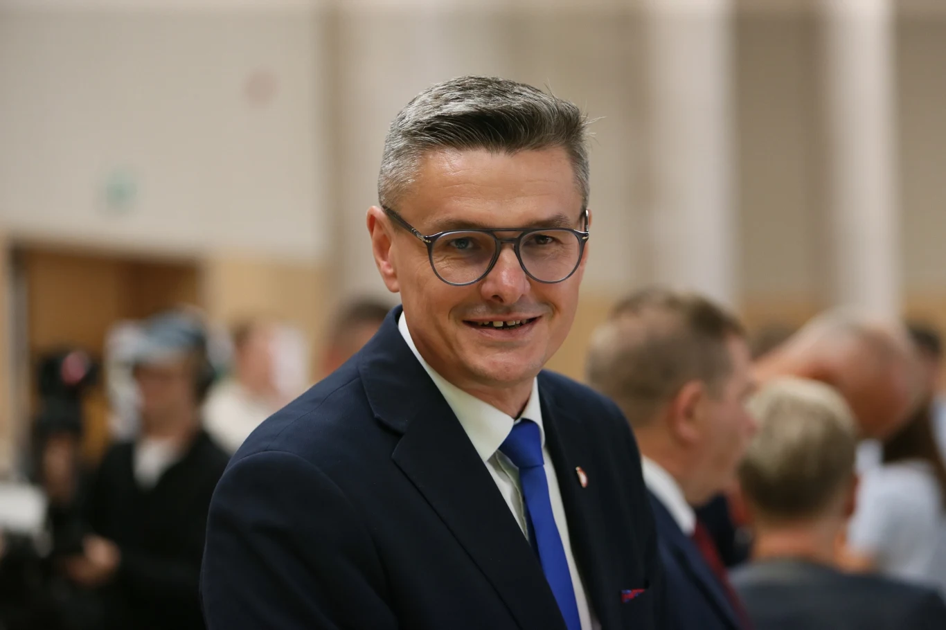 Nowo wybrany marszałek województwa lubuskiego Sebastian Ciemnoczołowski Mężczyzna w garniturze i okularach z uśmiechem na twarzy na tle nieostrego tłumu osób na sali, w formalnym otoczeniu.