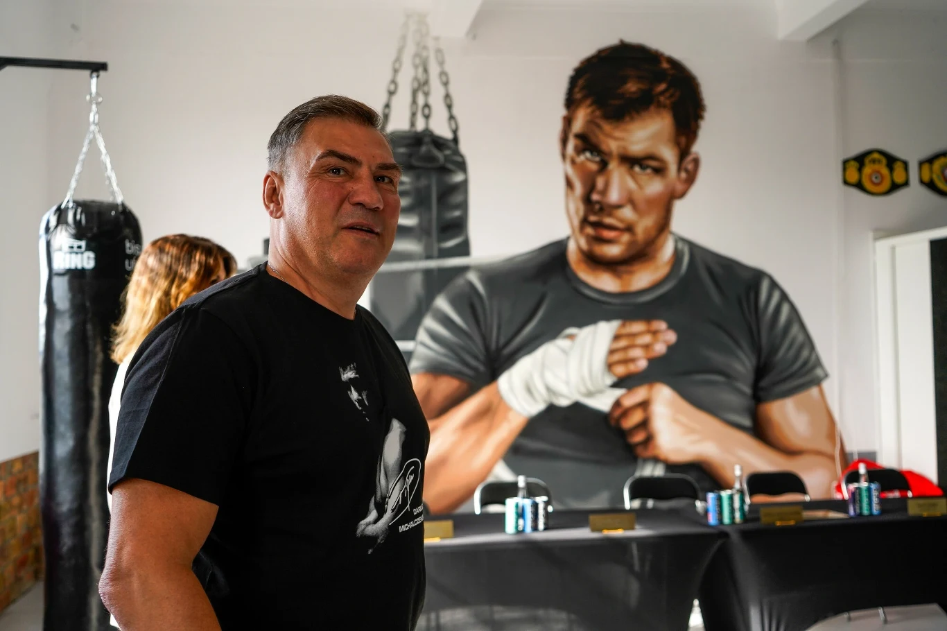 Mężczyzna w czarnej koszulce stoi w sali treningowej do boksu, w tle widoczny jest duży mural przedstawiający boksera, obok wiszą worki treningowe i na stole ustawione są napoje oraz pasy mistrzowskie.