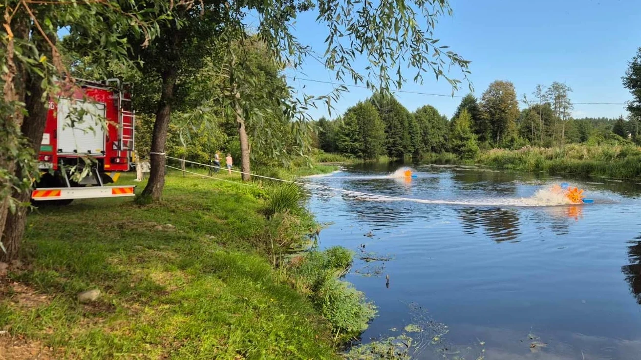 Śnięte ryby we Wkrze. Wyłowiono już 830 kg Wóz strażacki ustawiony na brzegu rzeki, kilka osób w żółtych kamizelkach ratunkowych uczestniczy w akcji na wodzie, na powierzchni widoczne są czerwone boje, dookoła zieleń i drzewa.