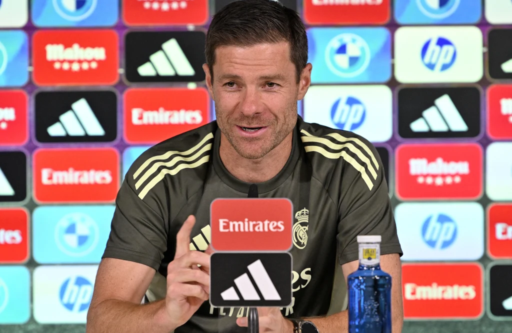 Mężczyzna w sportowej koszulce Realu Madryt siedzi przy stole konferencyjnym, odpowiada na pytania podczas konferencji prasowej, za nim tło z logotypami sponsorów.