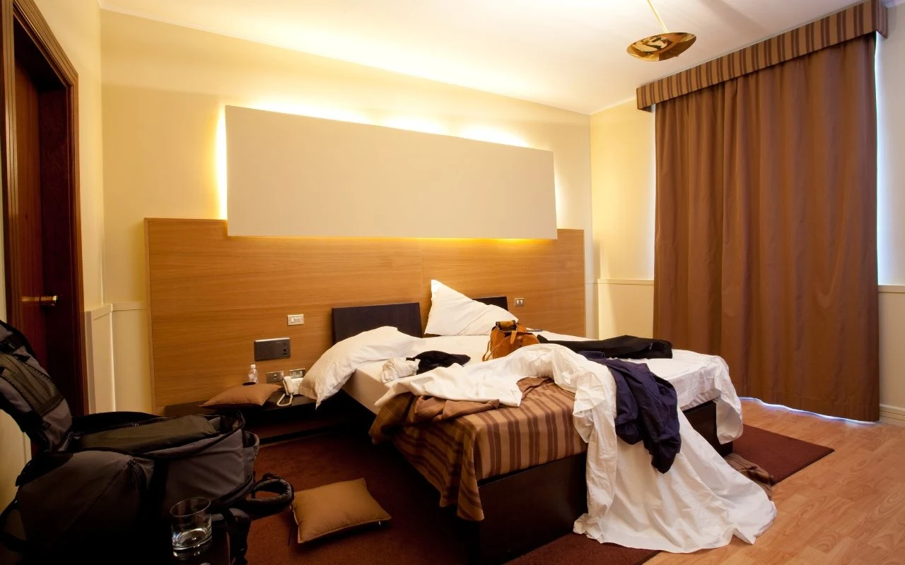 Standard hotelu często bywa dużym rozczarowaniem przy wyborze tanich ofert wakacji Rozrzucona pościel i ubrania na dwóch połączonych łóżkach w hotelowym pokoju, obok stoją torba podróżna i plecak, na łóżku leżą akcesoria osobiste, na podłodze poduszka, zasłony są zasłonięte.