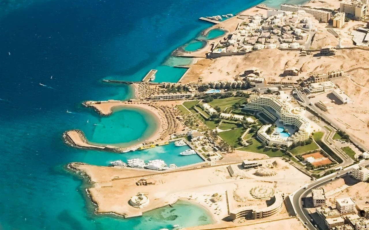 Hurghada to popularny kierunek wśród ofert "last minute" Widok z lotu ptaka na nadmorskie miasto z licznymi hotelami, basenami i prywatnymi plażami otoczonymi lazurową wodą oraz przystanią z zacumowanymi jachtami, otoczone pustynnym terenem.