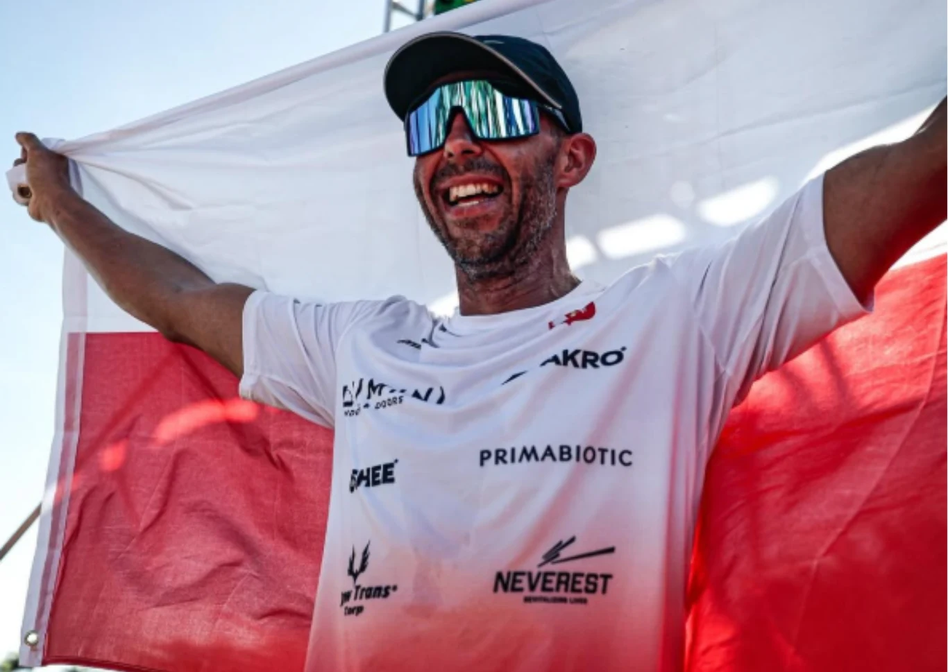 Jurand Czabański Sportowiec w białej koszulce z nadrukami sponsorów trzyma biało-czerwoną flagę Polski, uśmiechnięty, w okularach przeciwsłonecznych, ukazujący radość i dumę z osiągnięcia.