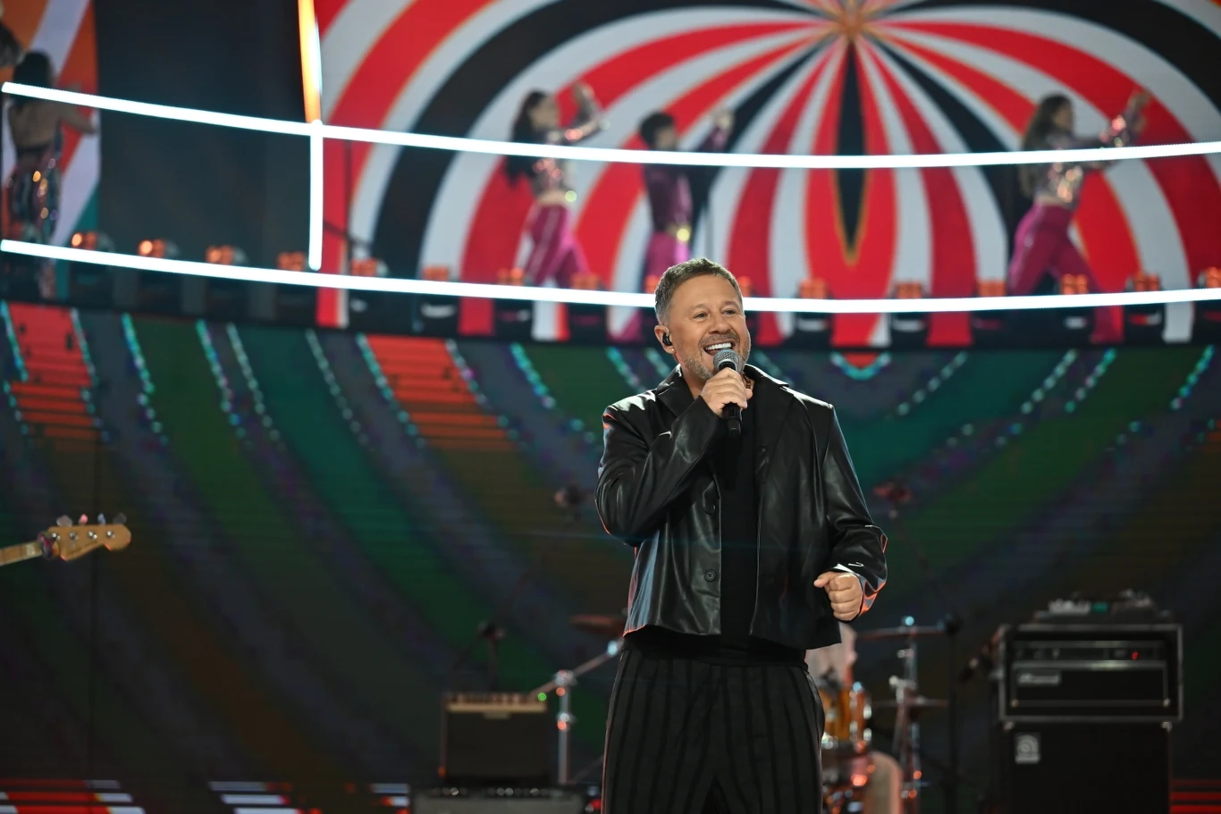 Top of the Top Sopot Festival 2025: Andrzej Piaseczny Mężczyzna w czarnej kurtce śpiewa na scenie z mikrofonem, w tle kolorowa scenografia z geometrycznym wzorem, zespół muzyczny oraz tancerki na podwyższeniu.