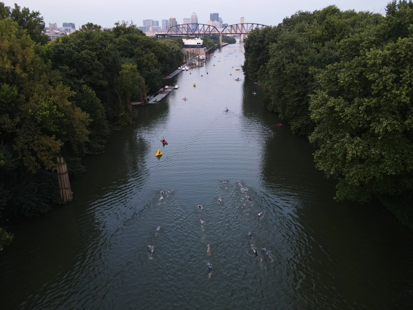 Sportowcy biorący udział w ironmanie Grupa osób płynie wzdłuż szerokiej rzeki otoczonej gęstymi drzewami, w oddali widoczny most oraz panorama miasta.
