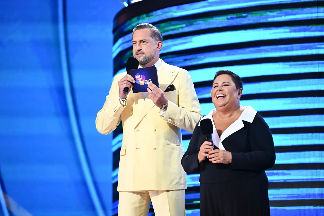 Dorota Wellman i Marcin Prokop prowadzą pierwszy wieczór festiwalu w Sopocie Dwóch prowadzących na scenie podczas wydarzenia, mężczyzna w jasnym garniturze trzyma kartkę z logotypem, kobieta ubrana na czarno trzyma mikrofon i szeroko się uśmiecha, w tle jasnoniebieskie światła sceniczne.