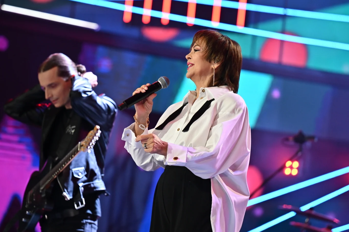 Izabela Trojanowska wystąpiła w Sopocie Kobieta w białej koszuli śpiewa do mikrofonu, obok niej stoi gitarzysta z długimi włosami, na scenie kolorowe światła i dynamiczne, neonowe tło.