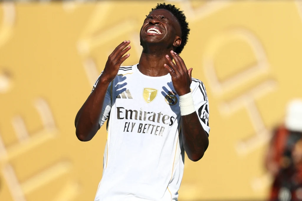 Piłkarz w białym stroju sportowym z logo Realu Madryt z uniesionymi rękami i wyrazem frustracji na twarzy, rozmyte złote tło.