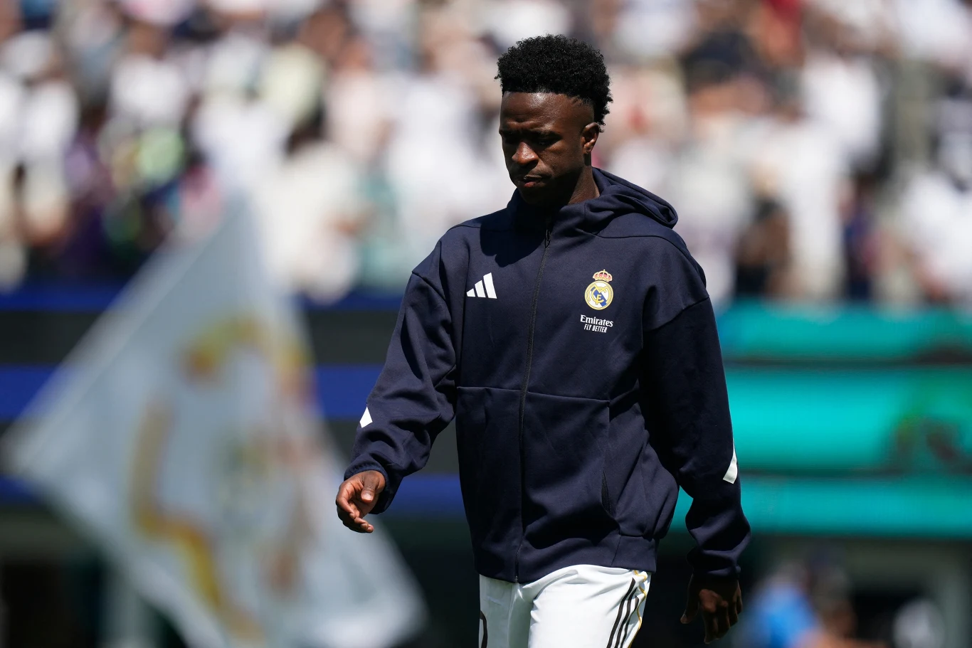 Młody piłkarz w ciemnej kurtce sportowej z herbem Realu Madryt idzie po boisku, w tle rozmyta flaga klubowa oraz kibice na trybunach.