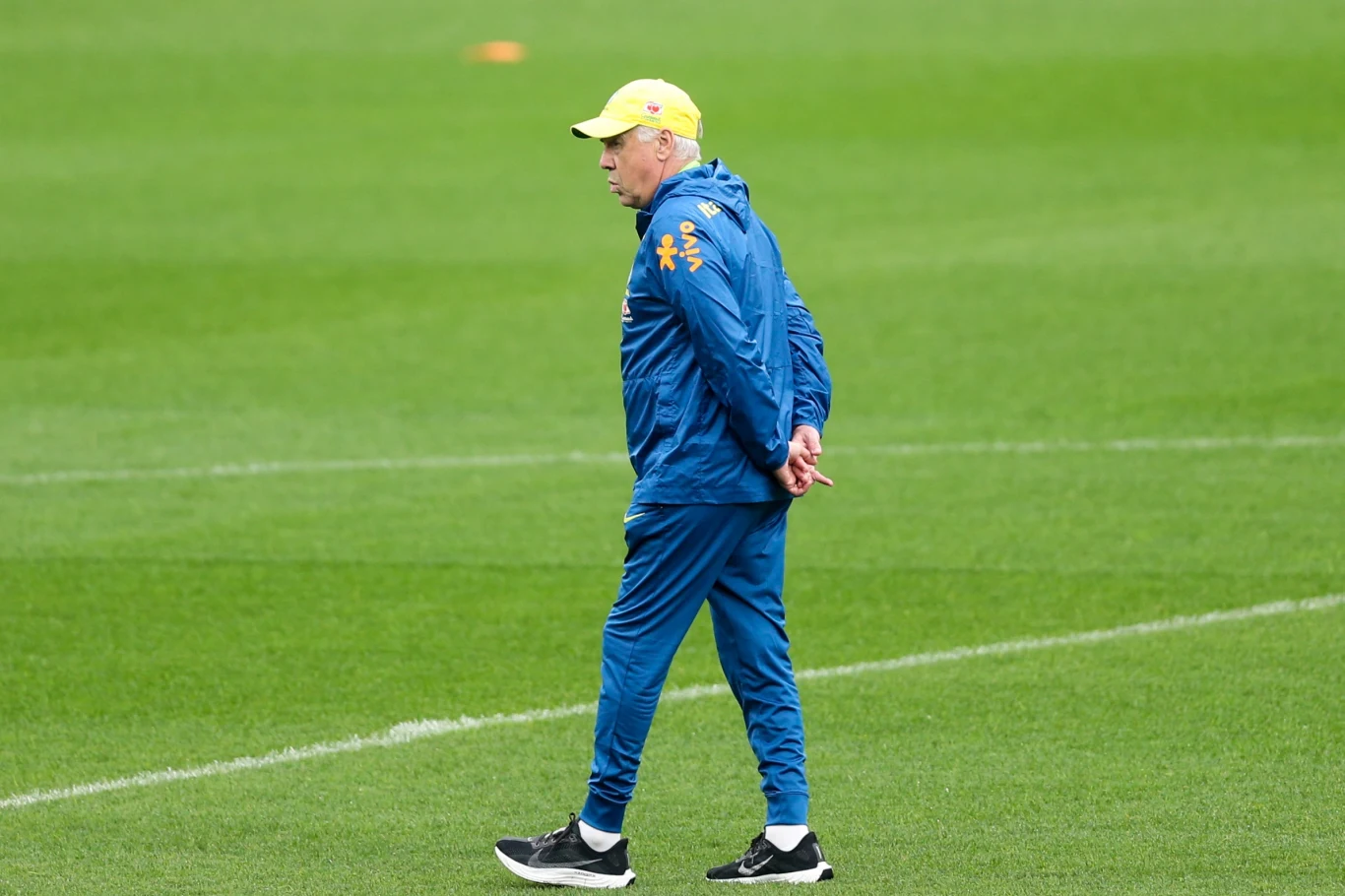 Mężczyzna w żółtej czapce oraz niebieskim dresie spaceruje po murawie boiska sportowego, trzymając ręce za plecami.