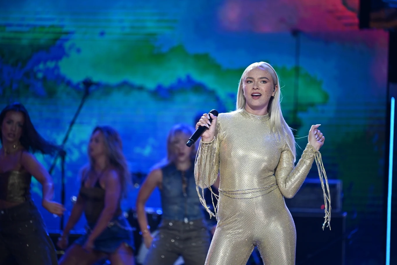 Natalia Nykiel podczas festiwalu w Sopocie 2025 Wokalistka w złotym kostiumie śpiewa na scenie, w tle grupa tancerek oraz kolorowa, abstrakcyjna grafika oświetlająca scenę.