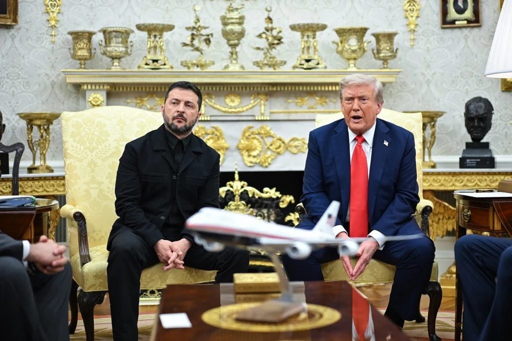 Dwóch mężczyzn siedzących na fotelach w wystawnie urządzonym pomieszczeniu z kominkiem i złotymi dekoracjami, na pierwszym planie stoi model samolotu typu passenger jet.