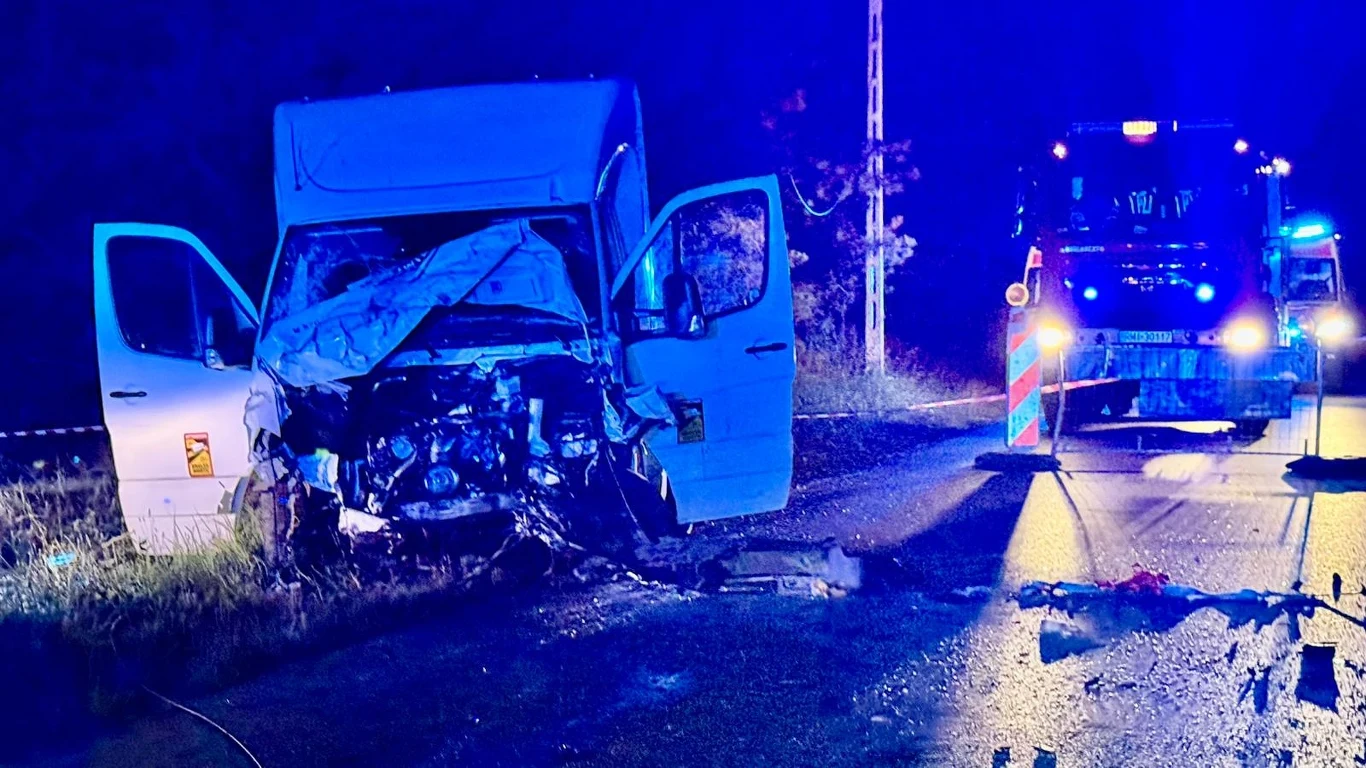 Wypadek w Grochowem. Zginęły trzy młode osoby Zniszczony przód białego samochodu dostawczego stojącego na poboczu, otwarte drzwi, ślady uszkodzeń na jezdni oraz pojazd służb ratowniczych z włączonymi światłami ostrzegawczymi w tle.
