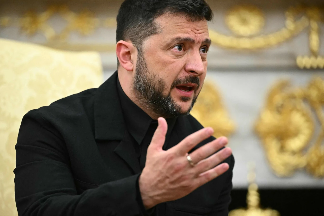 Mężczyzna z brodą i krótkimi włosami w czarnym ubraniu gestykuluje podczas rozmowy, w tle złote dekoracje i kawałek kremowego fotela.