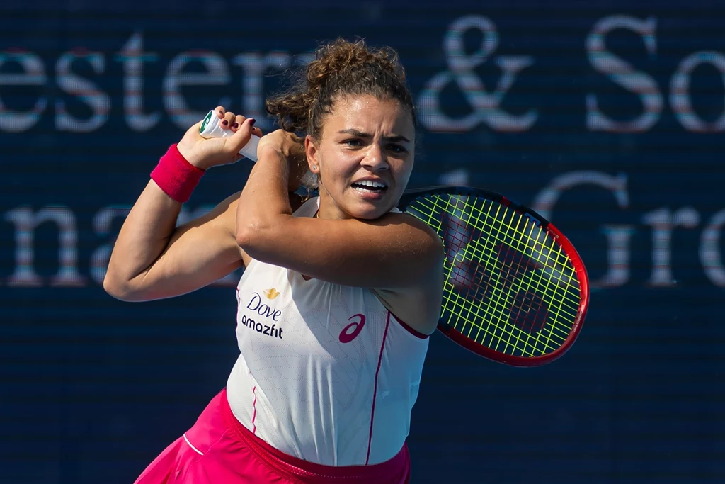 Jasmine Paolini Tenisistka w dynamicznej pozie, trzymająca rakietę tenisową po wykonaniu uderzenia, ubrana w sportowy strój z logo sponsora na tle rozmytego, ciemnego tła.