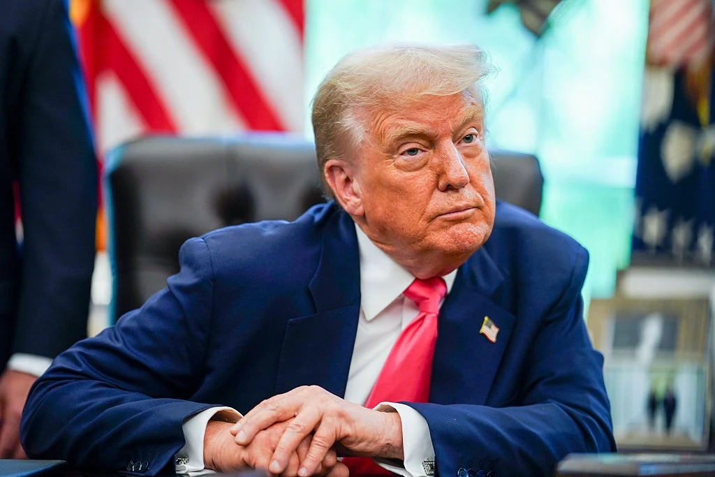 Amerykanie wyrazili swoje poparcie dla Donalda Trumpa. Jest najnowszy sondaż Mężczyzna w średnim wieku w garniturze z czerwonym krawatem siedzi przy biurku. W tle widoczne są flagi Stanów Zjednoczonych, a całość otoczenia sugeruje formalny kontekst biurowy lub urzędowy.