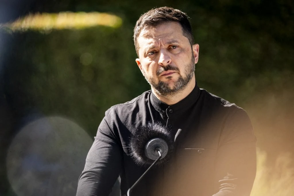 Mężczyzna w ciemnej koszuli, stojący przed mikrofonem na tle rozmytej zieleni, patrzący poważnie w stronę aparatu.