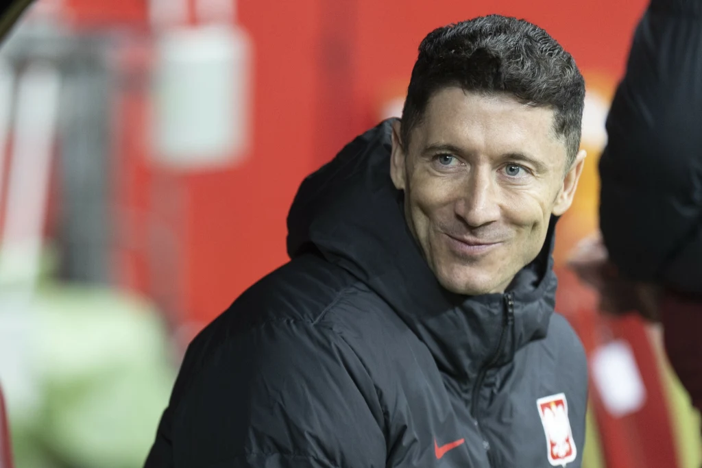 Robert Lewandowski Mężczyzna w ciepłej sportowej kurtce z godłem Polski na piersi uśmiecha się, siedząc w jasnym otoczeniu stadionowym z rozmytym tłem.