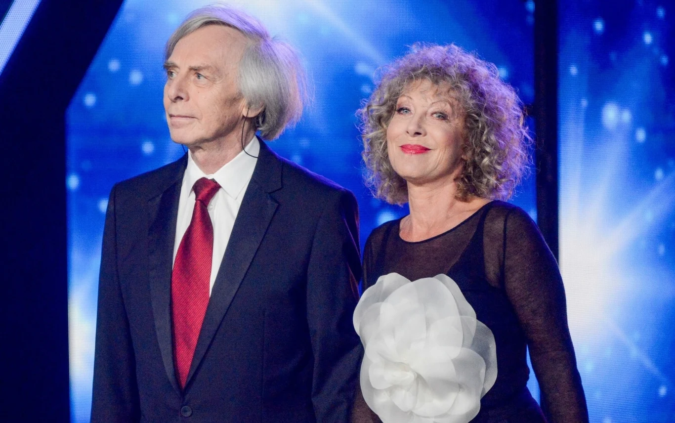 Włodzimierz Korcz i Alicja Majewska są na scenie od 50 lat Elegancko ubrany mężczyzna w ciemnym garniturze z czerwonym krawatem oraz kobieta z kręconymi włosami w czarnej sukni z dużą białą aplikacją kwiatową stoją razem na tle rozświetlonej sceny.