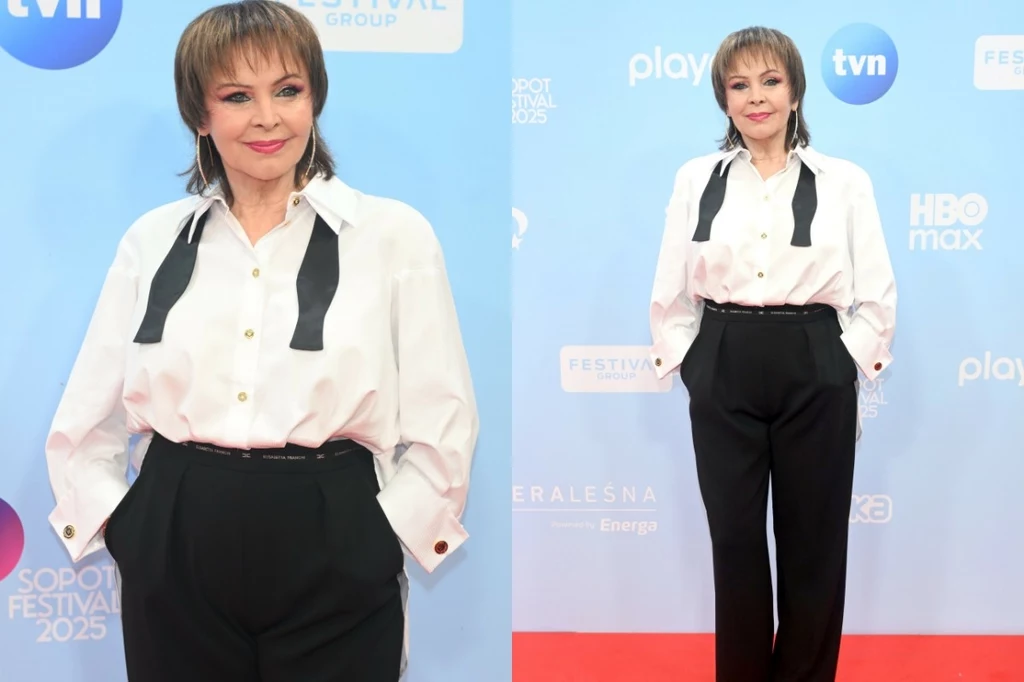 Elegancko ubrana kobieta w białej koszuli z rozpiętym krawatem oraz czarnych spodniach pozuje na tle ścianki z logotypami znanych stacji telewizyjnych i nazwą festiwalu Sopot Festival 2025, stojąc na czerwonym dywanie.