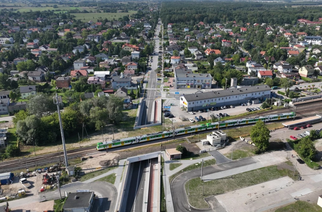 Widok miasta z lotu ptaka z dworcem kolejowym, przejazdem podziemnym dla samochodów oraz jadącym pociągiem, otoczonego licznymi domami, budynkami usługowymi i zielenią.