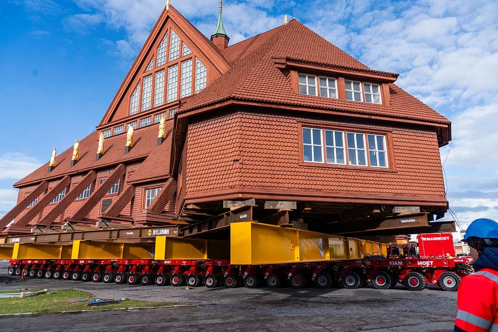 Zabytkowy kościół w Kirunie zostanie przeniesiony w inne miejsce na ogromnych platformach Duży drewniany budynek o stromym dachu transportowany przez specjalistyczną platformę z wieloma kołami, widocznych kilku pracowników w kaskach i kamizelkach ochronnych nadzorujących operację.